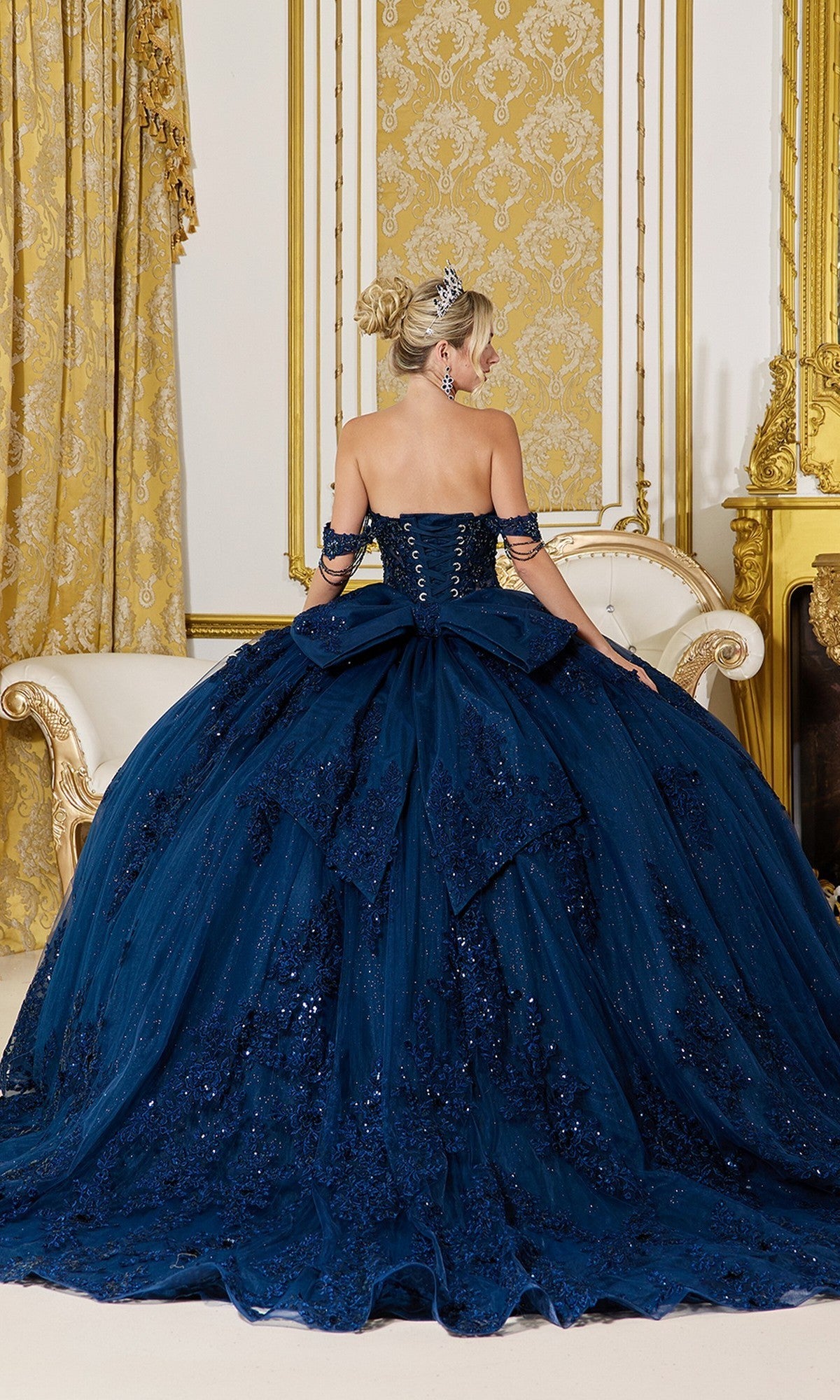 A woman in the Quinceanera Dress 5141 by Dancing Queen, featuring navy blue sequins and a full skirt, sits on a cream sofa in an ornate gold-accented room, facing away from the camera.